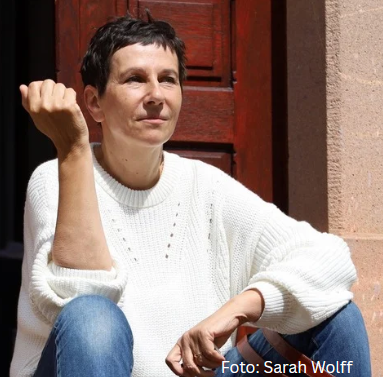 Frau mit kurzen Haaren und weißem Pullover, sitzend vor einer Tür